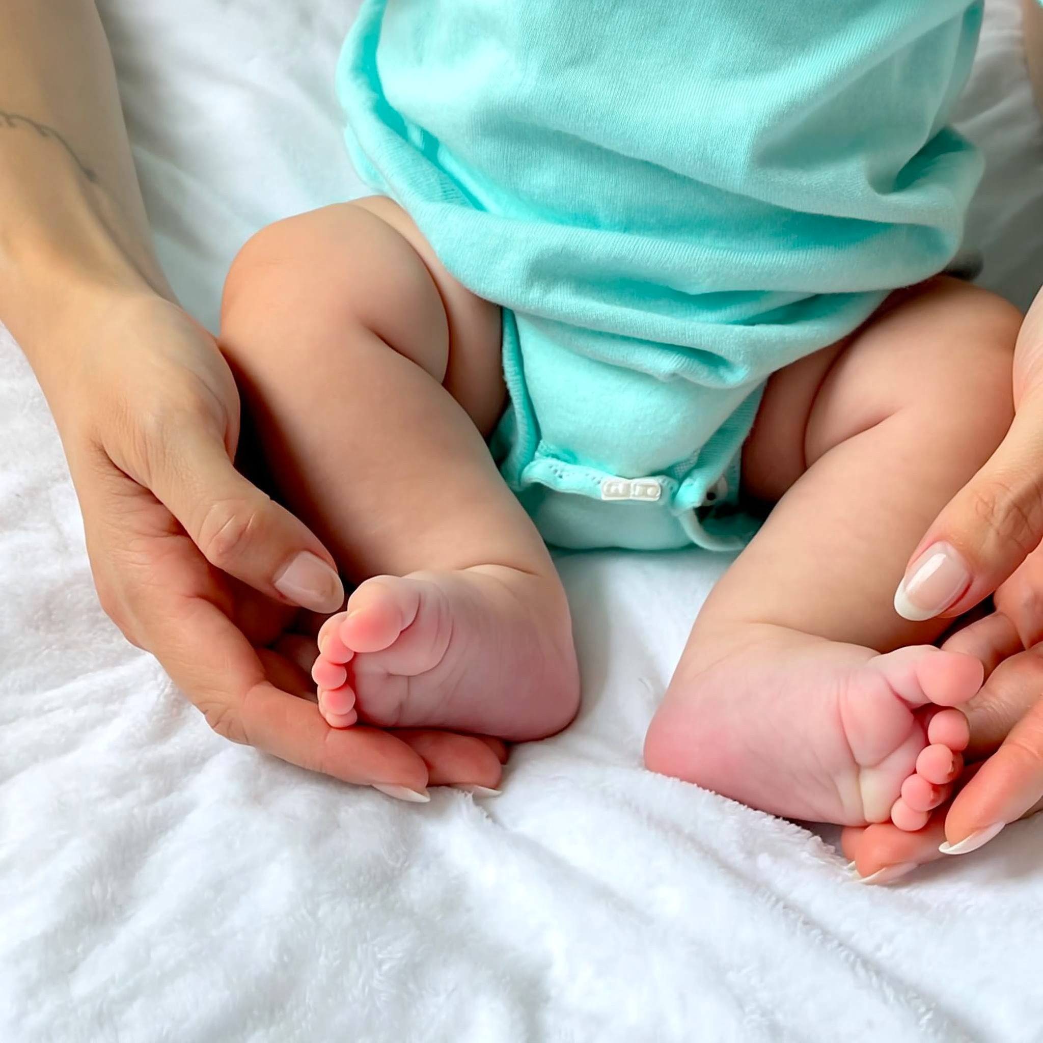 Twee handen houden de kleine voetjes van een baby in een turquoise rompertje vast